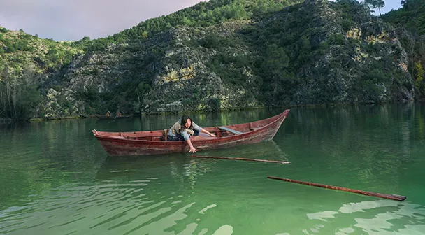 Eine Frau befindet sich mit einem Ruderboot aus Holz auf einem See und bückt sich nach den Paddeln, die aus dem Boot gefallen sind.